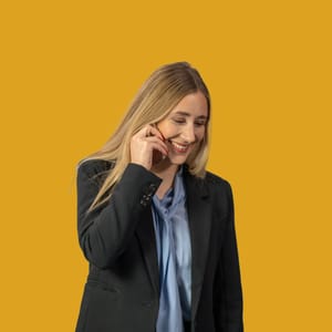 Frau in schwarzem Anzug telefoniert und lächelt vor gelbem Hintergrund.