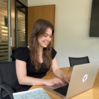 Frau sitzt am Tisch und arbeitet lächelnd an einem Laptop.