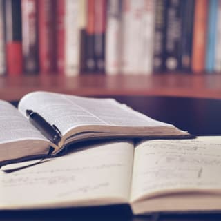 Offene Bücher mit Stift auf einem Tisch vor einem Bücherregal.