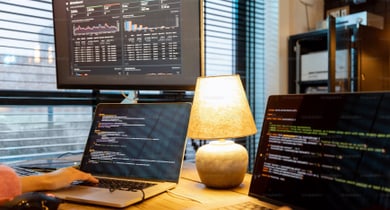 Person am Schreibtisch mit Laptops und Bildschirm voller Code.