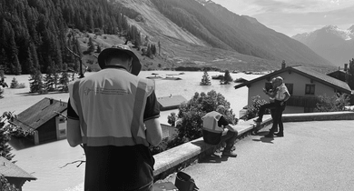 Drei Personen bei der Arbeit in bergiger Landschaft mit Häusern.