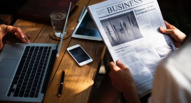 Eine Person liest eine Businesszeitung während eine andere Person am Laptop arbeitet.