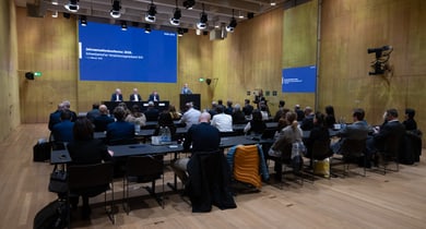 Konferenzraum mit Rednern und Publikum auf Stühlen.