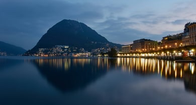 Lugano bei Dämmerung über einem ruhigen See mit Lichtern entlang des Ufers.