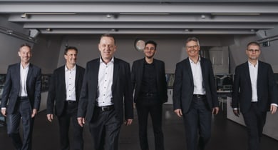 Sechs Männer in Anzügen gehen auf die Kamera zu.