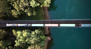Zug fährt über Brücke mit Bäumen und Fluss darunter, Luftansicht.