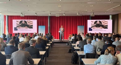 Konferenzraum mit vielen Menschen und einer Person auf der Bühne.