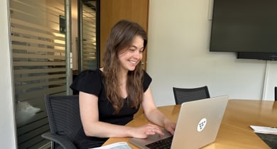 Frau sitzt am Tisch und arbeitet lächelnd an einem Laptop.