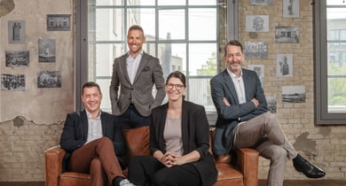 Vier Personen sitzen und stehen in einem Raum mit Backsteinmauern und Bildern.