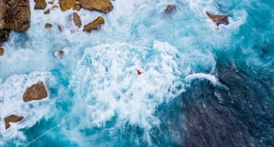Luftbild einer Person in rauer Brandung neben Felsen.