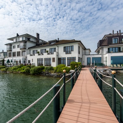 Steg führt zu Häusern am Seeufer mit bewölktem Himmel.