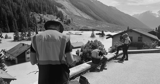 Drei Personen bei der Arbeit in bergiger Landschaft mit Häusern.