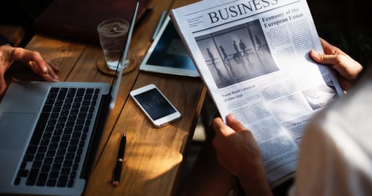 Eine Person liest eine Businesszeitung während eine andere Person am Laptop arbeitet.