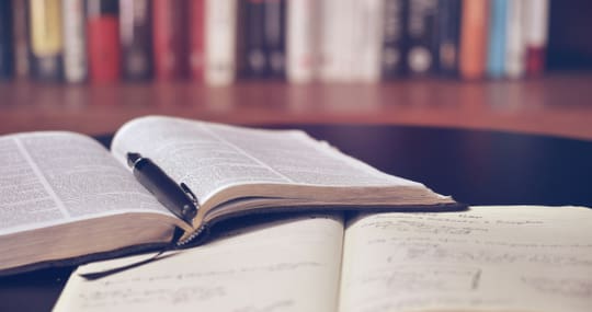 Offene Bücher mit Stift auf einem Tisch vor einem Bücherregal.
