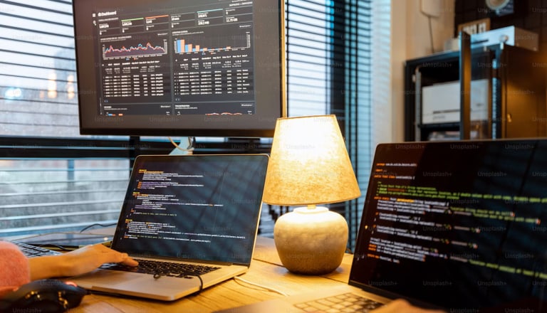 Person am Schreibtisch mit Laptops und Bildschirm voller Code.
