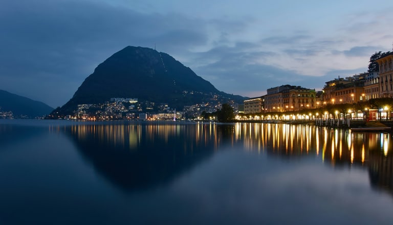 Lugano bei Dämmerung über einem ruhigen See mit Lichtern entlang des Ufers.