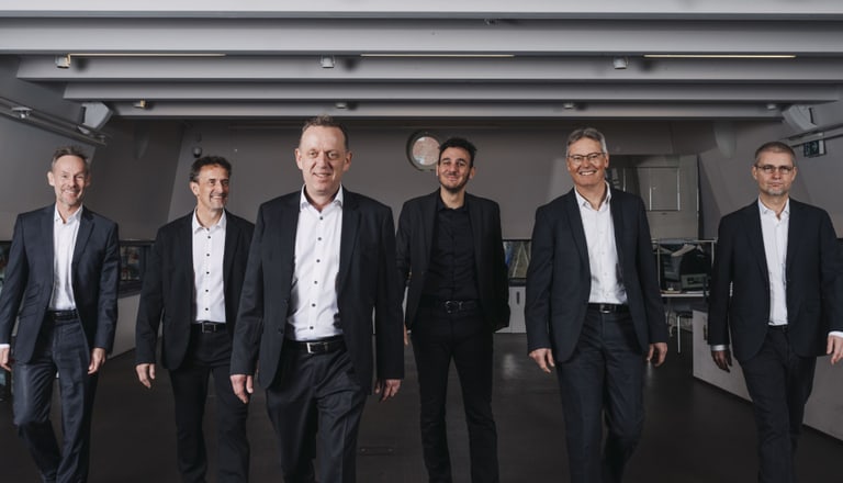 Sechs Männer in Anzügen gehen auf die Kamera zu.