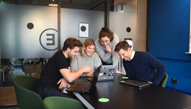 Vier Personen an einem Tisch, die auf einen Laptop schauen und lächeln.