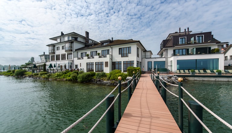 Steg führt zu Häusern am Seeufer mit bewölktem Himmel.