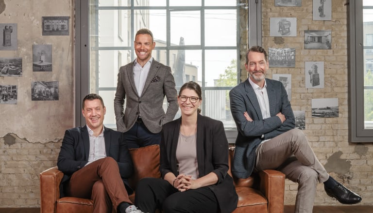 Vier Personen sitzen und stehen in einem Raum mit Backsteinmauern und Bildern.