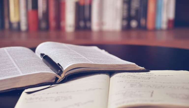 Offene Bücher mit Stift auf einem Tisch vor einem Bücherregal.