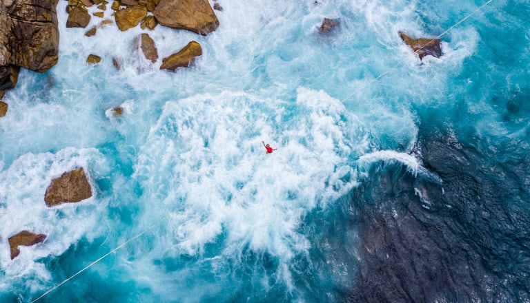 Luftbild einer Person in rauer Brandung neben Felsen.