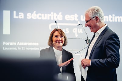 Zwei Personen lächelnd bei einer Veranstaltung mit Präsentationsbildschirm im Hintergrund.