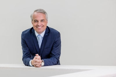 Person in blauem Anzug lächelt und lehnt sich auf weisse Barriere.