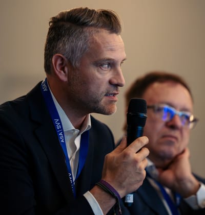 Homme avec un micro parle lors d'un événement, deuxième homme en arrière-plan.