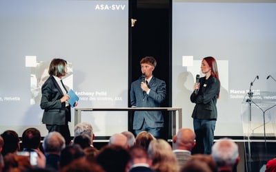 Drei Personen sprechen bei einer Konferenz auf der Bühne, Publikum im Vordergrund.