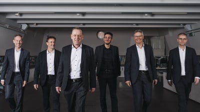 Sechs Männer in Anzügen gehen auf die Kamera zu.