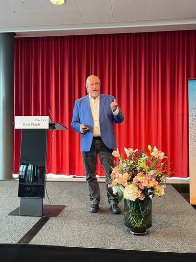 Eine Person in einem blauen Blazer steht auf einer Bühne vor einem roten Vorhang.